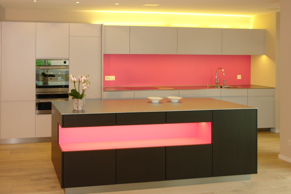 Grosszügige, weisse Küchenschränke und schöner, schwarzer Kochinsel in der Mitte. Beides mit beleuchteten Aussparungen in Pink für den Farbklecks. Ihr Küchenwunsch wird wahr mit der Firma Weidmann AG in Möhlin.